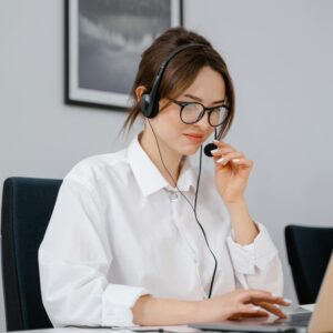 Young woman in glasses and headset providing customer support at a laptop in an office setting.